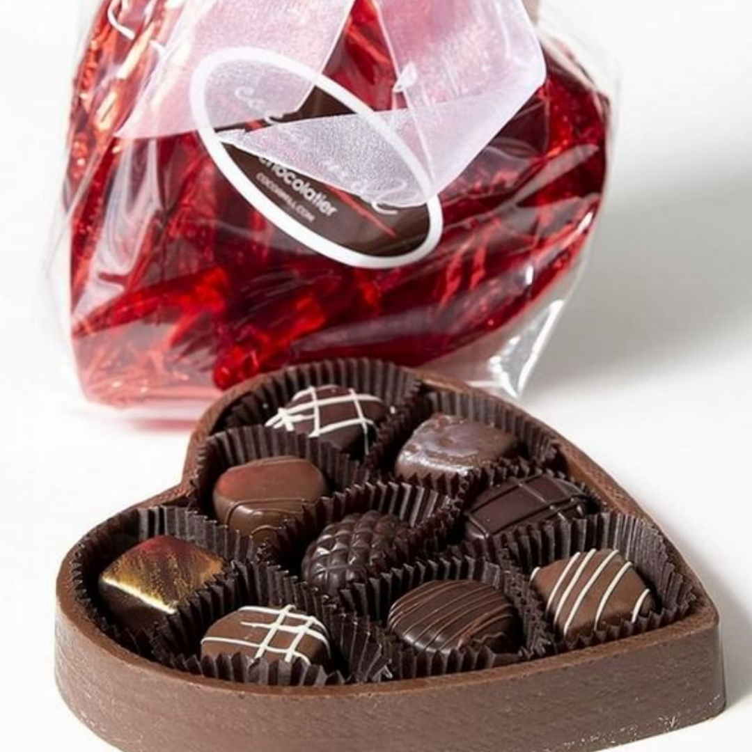 A heart-shaped box made from chocolate that contains nine truffles in black candy papers with a red lid and cellophane behind it