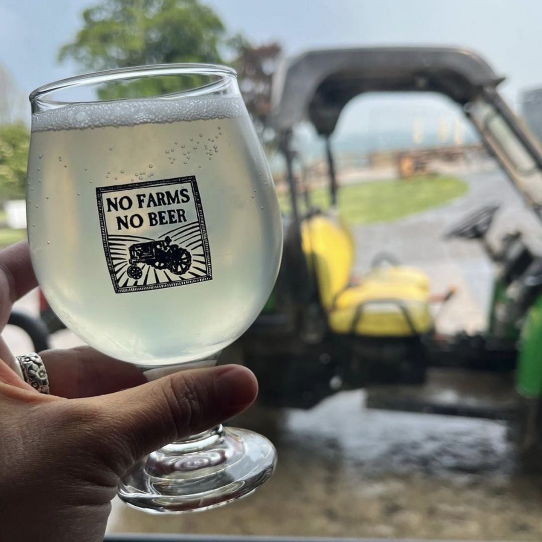 A hand holding a glass of beer in front of a tractor. The glass has a graphic image of a tractor with the words “No Farms No Beer”