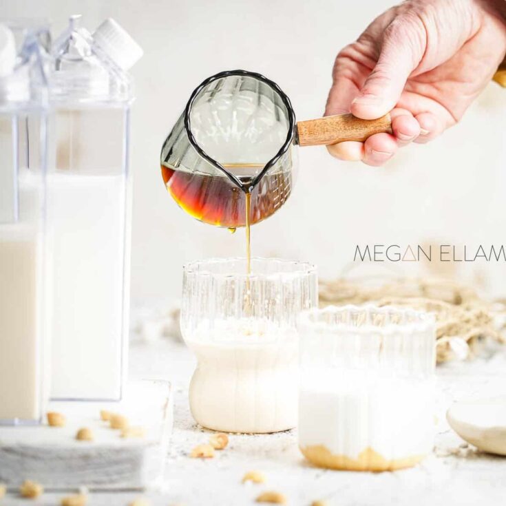 Sweet vanilla syrup being poured into a glass, with bottles of vanilla extract and ingredients in th.