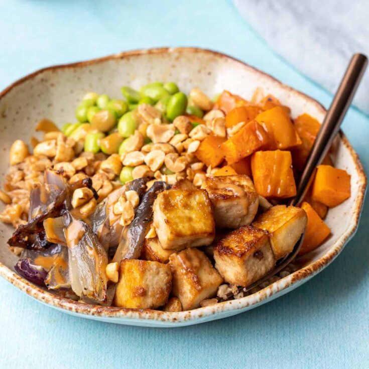 Savory tofu and vegetable stir-fry with sweet potatoes, edamame, and peanuts in a ceramic bowl.