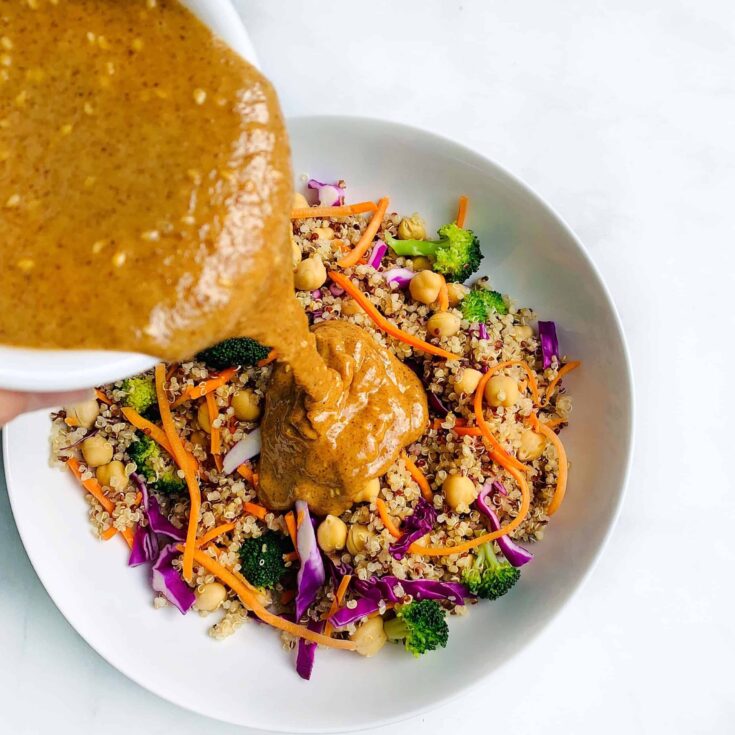 Creamy peanut sauce being poured over a colorful vegetable quinoa salad with broccoli, carrots, and.